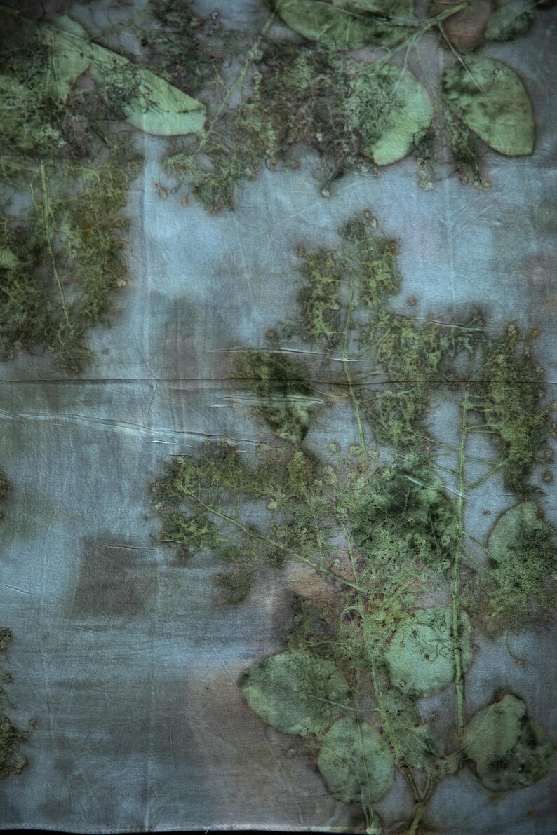 May include: A piece of fabric featuring a botanical print with green leaf and branch imprints on a muted blue and grey background. The textile displays organic, textured patterns created by the natural shapes of foliage pressed into the material.