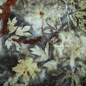 May include: A textile piece featuring a botanical print with dark gray and black background tones. The surface displays detailed imprints of various leaves and flowers in shades of cream, pale green, and burnt orange, creating a layered organic pattern.