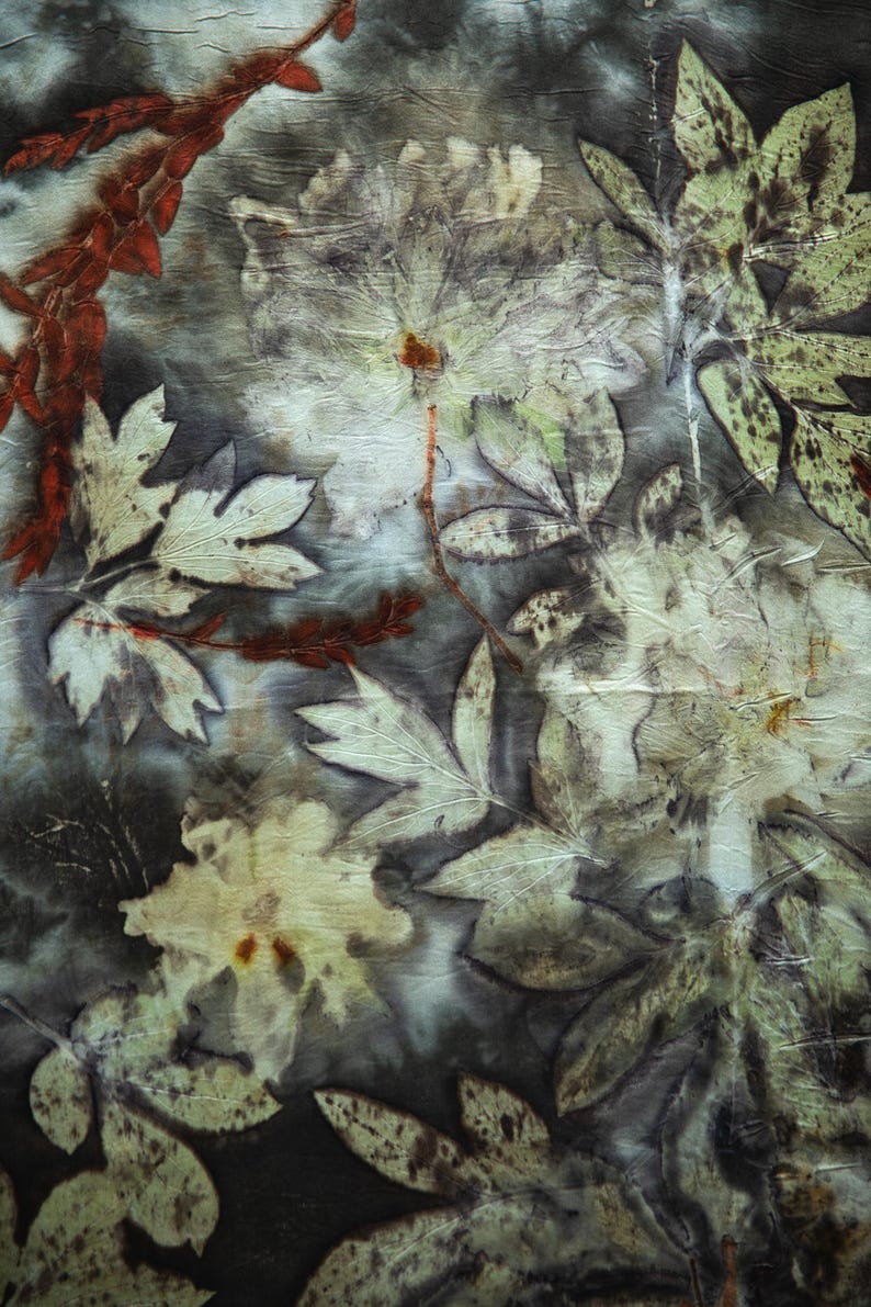 May include: A textile piece featuring a botanical print with dark gray and black background tones. The surface displays detailed imprints of various leaves and flowers in shades of cream, pale green, and burnt orange, creating a layered organic pattern.
