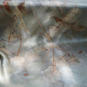 May include: A smooth, lustrous fabric featuring a botanical print with muted sage green and gray tones. The textile displays organic, rust-colored leaf and branch imprints scattered across the surface, creating a soft, layered visual effect.
