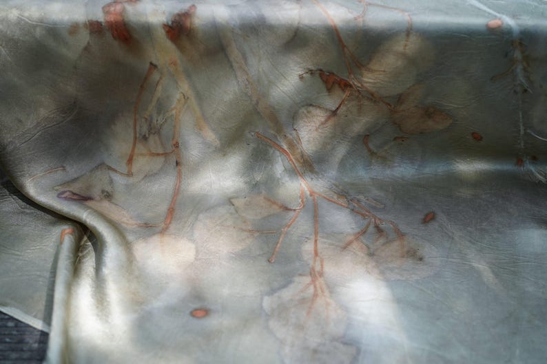 May include: A smooth, lustrous fabric featuring a botanical print with muted sage green and gray tones. The textile displays organic, rust-colored leaf and branch imprints scattered across the surface, creating a soft, layered visual effect.