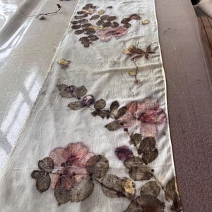 May include: A long, rectangular off-white fabric runner featuring a botanical print with pressed pink flowers, dark green leaves, and brown foliage. The textile has a crinkled texture and raw, unfinished edges, displaying a natural, organic pattern.