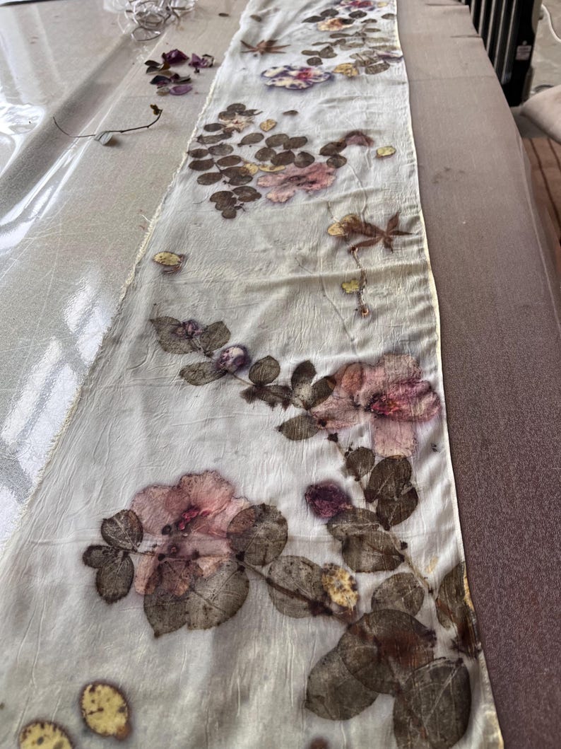 May include: A long, rectangular off-white fabric runner featuring a botanical print with pressed pink flowers, dark green leaves, and brown foliage. The textile has a crinkled texture and raw, unfinished edges, displaying a natural, organic pattern.