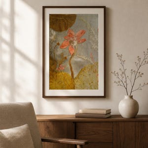 May include: A framed wall art print featuring a central coral-colored lotus flower with a long stem, surrounded by large, textured golden-brown lily pads and delicate white botanical elements against a soft, muted gray background.