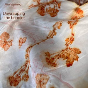 May include: A white fabric featuring botanical prints in shades of orange and rust, created through a steaming process. The text "After steaming Unwrapping the bundle" is printed in brown on the upper left side of the textile surface.