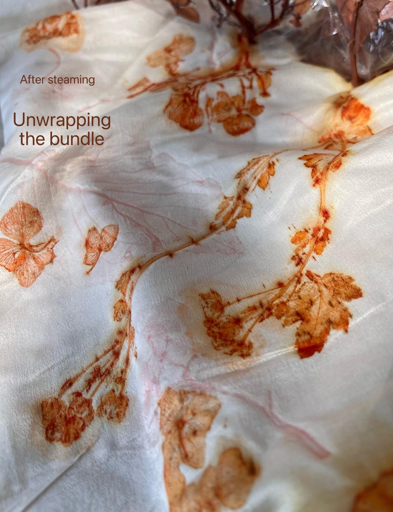 May include: A white fabric featuring botanical prints in shades of orange and rust, created through a steaming process. The text "After steaming Unwrapping the bundle" is printed in brown on the upper left side of the textile surface.