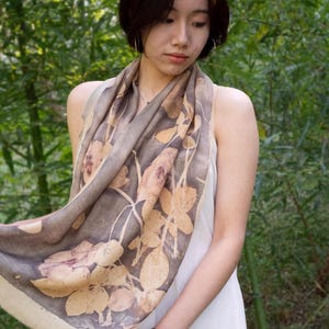 May include: A woman wearing a beige and brown floral print scarf over a white dress. The scarf features a botanical design with light brown and beige leaves and flowers. The background is a lush green bamboo forest.