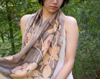 Puede incluir: Una mujer con una bufanda beige y marrón con estampado floral sobre un vestido blanco. La bufanda presenta un diseño botánico con hojas y flores de color marrón claro y beige. El fondo es un exuberante bosque de bambú verde.