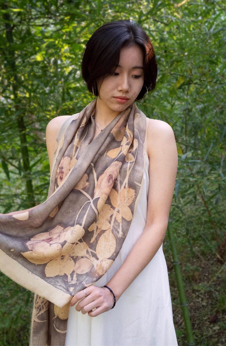 May include: A woman wearing a beige and brown floral print scarf over a white dress. The scarf features a botanical design with light brown and beige leaves and flowers. The background is a lush green bamboo forest.