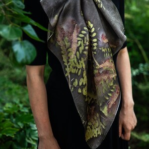 May include: A square scarf featuring a dark charcoal gray base with a botanical print of green and brown fern leaves and foliage. The scarf is tied around the neck and drapes over a solid black short-sleeved top, displaying intricate leaf patterns.