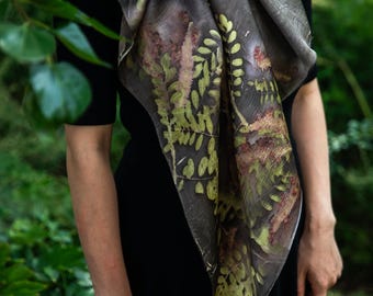 Puede incluir: Un pañuelo cuadrado con una base gris carbón oscuro y un estampado botánico de hojas de helecho y follaje en tonos verdes y marrones. El pañuelo está atado al cuello y cae sobre una blusa negra lisa de manga corta, mostrando patrones de hojas.