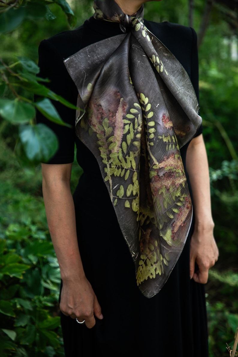 May include: A square scarf featuring a dark charcoal gray base with a botanical print of green and brown fern leaves and foliage. The scarf is tied around the neck and drapes over a solid black short-sleeved top, displaying intricate leaf patterns.