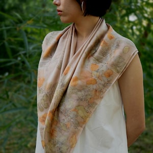 May include: A square scarf in a soft beige hue featuring a botanical print with scattered orange and muted green leaf patterns. The fabric drapes around the neck and shoulders, displaying a natural, organic design with detailed foliage imprints.