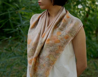 Puede incluir: Un pañuelo cuadrado en un tono beige suave con un estampado botánico de hojas dispersas en colores naranja y verde apagado. La tela se drapea alrededor del cuello y los hombros, mostrando un diseño natural y orgánico con impresiones detalladas de follaje.