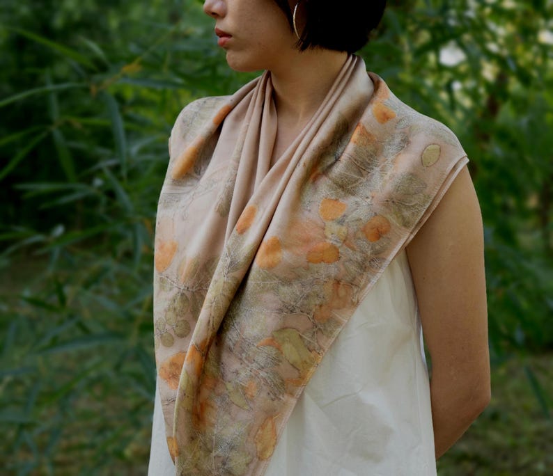 May include: A square scarf in a soft beige hue featuring a botanical print with scattered orange and muted green leaf patterns. The fabric drapes around the neck and shoulders, displaying a natural, organic design with detailed foliage imprints.