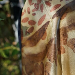 Pu&ograve; includere: Un foulard in tessuto color oro dalla consistenza liscia con una stampa botanica di foglie sparse nei toni del marrone e del ruggine. Il materiale presenta una lucentezza sottile e cade in pieghe morbide, mostrando un motivo ripetuto di sagome vegetali.