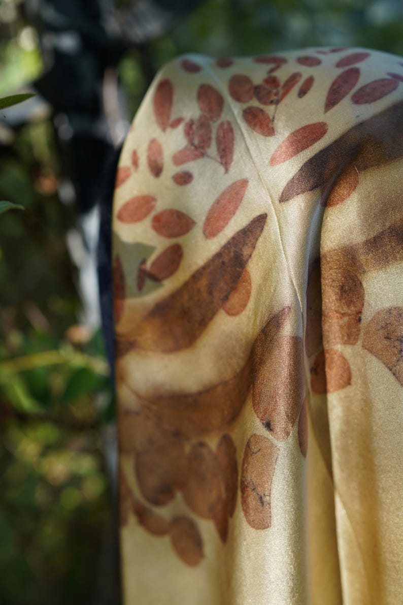 Pu&ograve; includere: Un foulard in tessuto color oro dalla consistenza liscia con una stampa botanica di foglie sparse nei toni del marrone e del ruggine. Il materiale presenta una lucentezza sottile e cade in pieghe morbide, mostrando un motivo ripetuto di sagome vegetali.