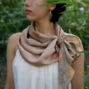 May include: A square silk scarf featuring a botanical print with muted earth tones, including shades of beige, soft brown, and faded orange. The scarf is tied in a knot at the side, displaying intricate leaf patterns and organic shapes across the fabric.