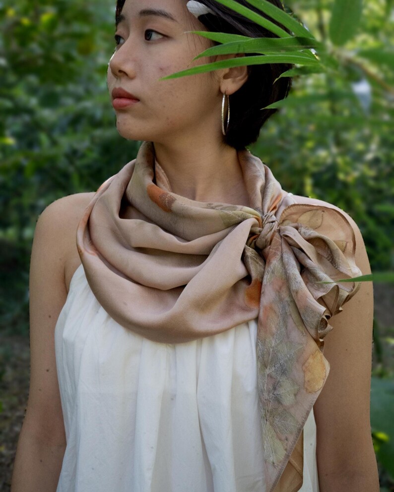 May include: A square silk scarf featuring a botanical print with muted earth tones, including shades of beige, soft brown, and faded orange. The scarf is tied in a knot at the side, displaying intricate leaf patterns and organic shapes across the fabric.