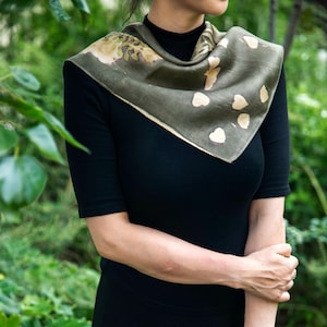 May include: A square scarf in a muted olive green hue featuring a botanical print of scattered cream-colored heart-shaped leaves and foliage patterns. The scarf is draped around the neck over a solid black short-sleeved top with a high neckline.