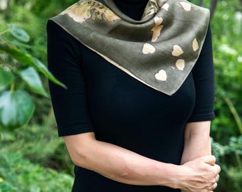 May include: A square scarf in a muted olive green hue featuring a botanical print of scattered cream-colored heart-shaped leaves and foliage patterns. The scarf is draped around the neck over a solid black short-sleeved top with a high neckline.