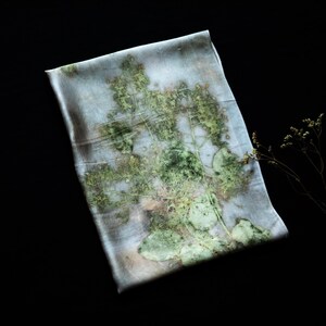 May include: A rectangular silk scarf featuring a botanical print with shades of pale blue, grey, and green. The fabric displays a detailed, organic pattern of pressed leaves and stems, creating a soft, layered visual effect across the surface.