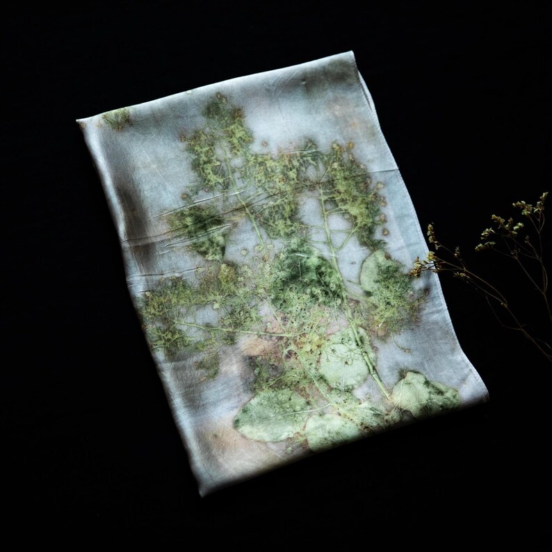May include: A rectangular silk scarf featuring a botanical print with shades of pale blue, grey, and green. The fabric displays a detailed, organic pattern of pressed leaves and stems, creating a soft, layered visual effect across the surface.