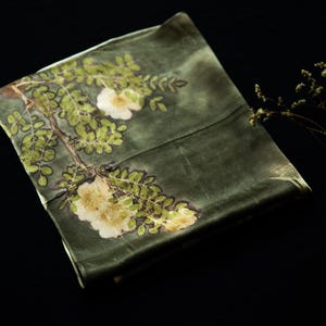 May include: A rectangular olive green fabric piece featuring a botanical print of white flowers and green leaves arranged along a branch. The textile has a smooth, slightly reflective surface and is displayed with a small dried plant sprig.