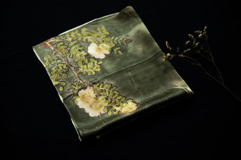 May include: A rectangular olive green fabric piece featuring a botanical print of white flowers and green leaves arranged along a branch. The textile has a smooth, slightly reflective surface and is displayed with a small dried plant sprig.