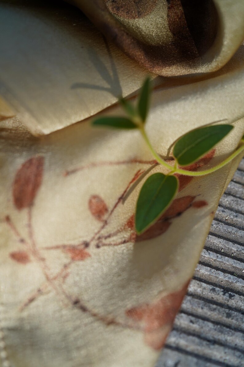 Pu&ograve; includere: Un tessuto beige chiaro con un motivo stampato di sagome di rami marroni con piccole foglie. Un piccolo rametto verde fresco con tre foglie appuntite poggia sulla superficie del tessuto, proiettando un'ombra distinta sul materiale.