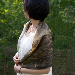 May include: A rectangular scarf featuring a dark brown and gold botanical print with a textured, leaf-like pattern. The scarf is draped over the shoulders, showcasing a smooth, lightweight fabric with a subtle sheen and deep, earthy color tones.