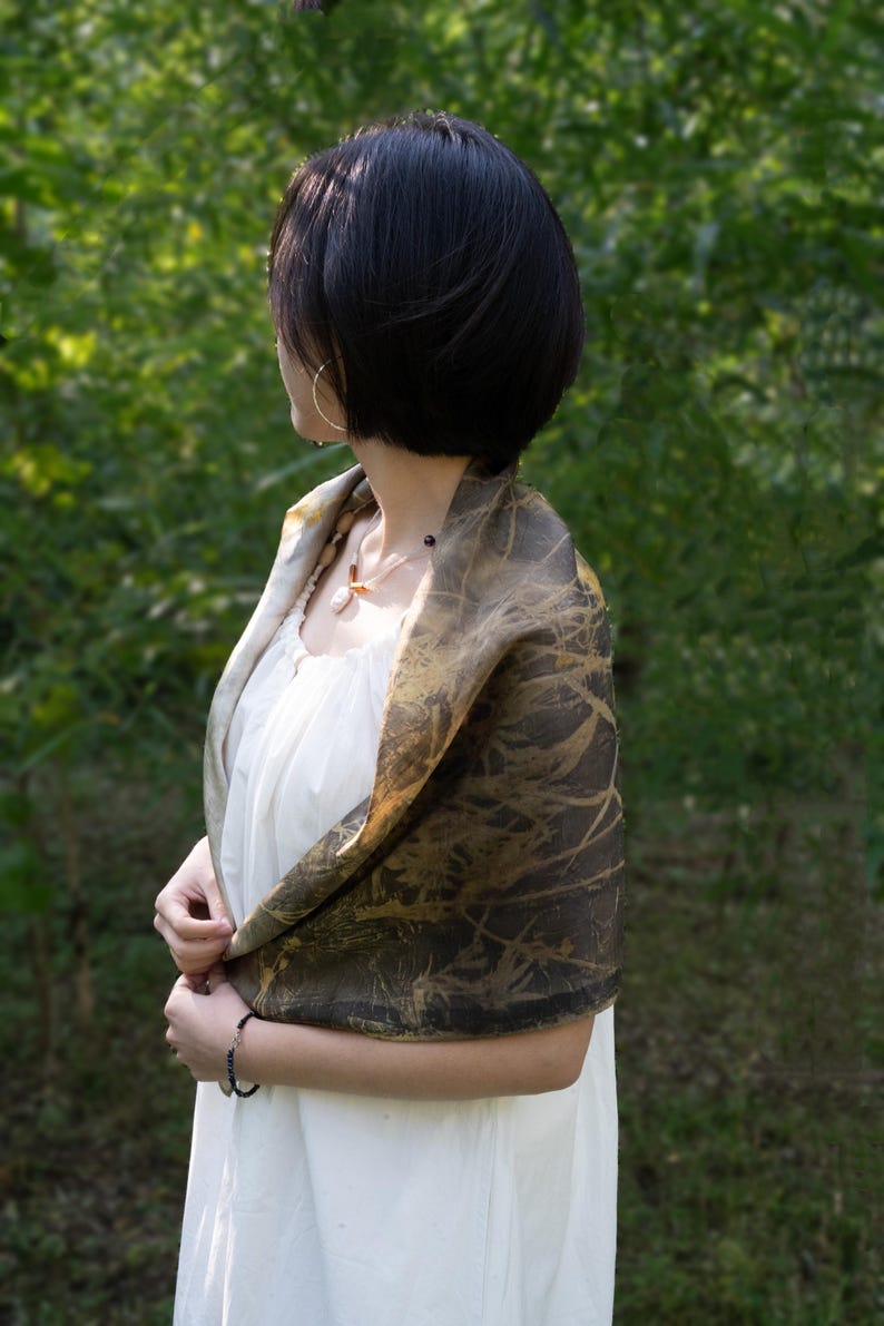 May include: A rectangular scarf featuring a dark brown and gold botanical print with a textured, leaf-like pattern. The scarf is draped over the shoulders, showcasing a smooth, lightweight fabric with a subtle sheen and deep, earthy color tones.