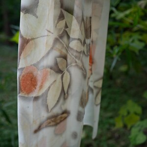 以下が含まれることがあります： 茶色、タン、オレンジ色の葉の植物柄がプリントされた、透け感のあるオフホワイトのスカーフ。緑の葉を背景に、屋外に吊るされています。生地は軽く、流れるように見えます。