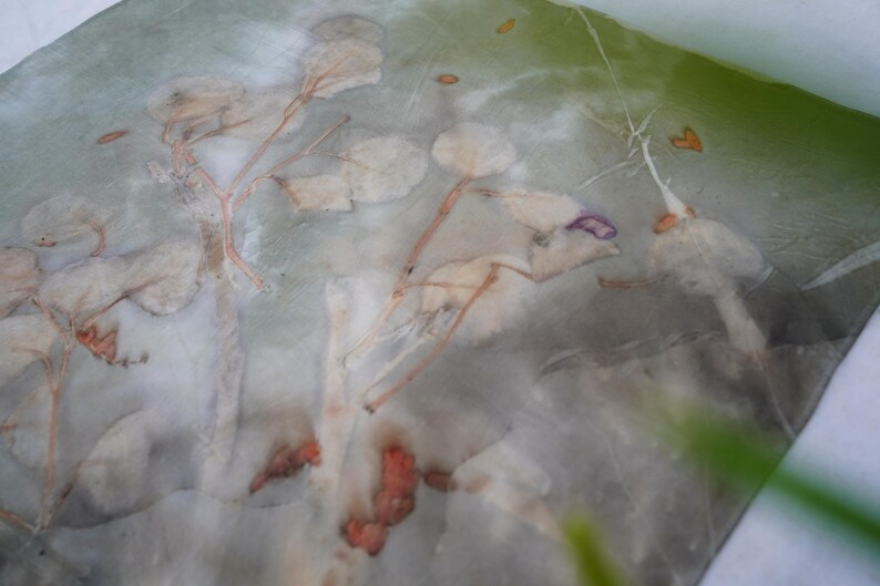 May include: A semi-sheer fabric featuring a botanical print with muted green, gray, and white tones. The surface displays organic leaf and stem patterns in soft, faded shades of orange, brown, and purple, creating a layered, natural appearance.