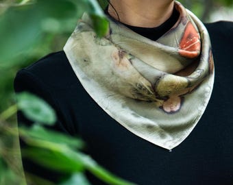 Puede incluir: Un pañuelo cuadrado de seda con un estampado botánico en tonos verdes apagados, grises y naranjas terrosos. El pañuelo está doblado en forma triangular y se lleva alrededor del cuello sobre una prenda negra de manga larga, mostrando patrones de hojas.