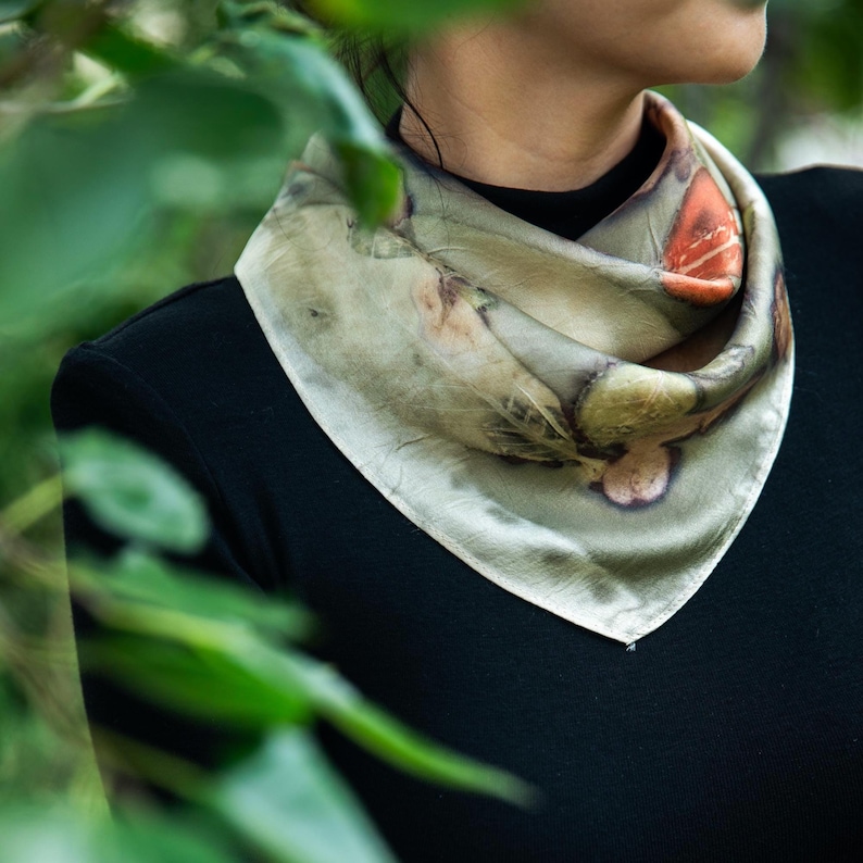 May include: A square silk scarf featuring a botanical print with muted green, grey, and earthy orange tones. The scarf is folded into a triangular shape and worn around the neck over a solid black long-sleeved garment, displaying organic leaf patterns.