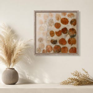 May include: A square wall art piece in a light wood frame featuring a grid of pressed, circular orange and beige leaves on a textured off-white background. The artwork displays varying shades of rust, copper, and cream in a repeating pattern.