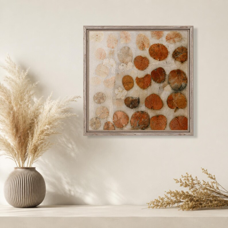 May include: A square wall art piece in a light wood frame featuring a grid of pressed, circular orange and beige leaves on a textured off-white background. The artwork displays varying shades of rust, copper, and cream in a repeating pattern.