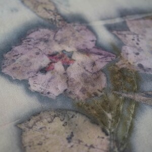 May include: Close-up of a textile with a floral print. The design features a large, faded purple flower with a red center and several leaves in shades of brown and green. The fabric has a soft, muted color palette.