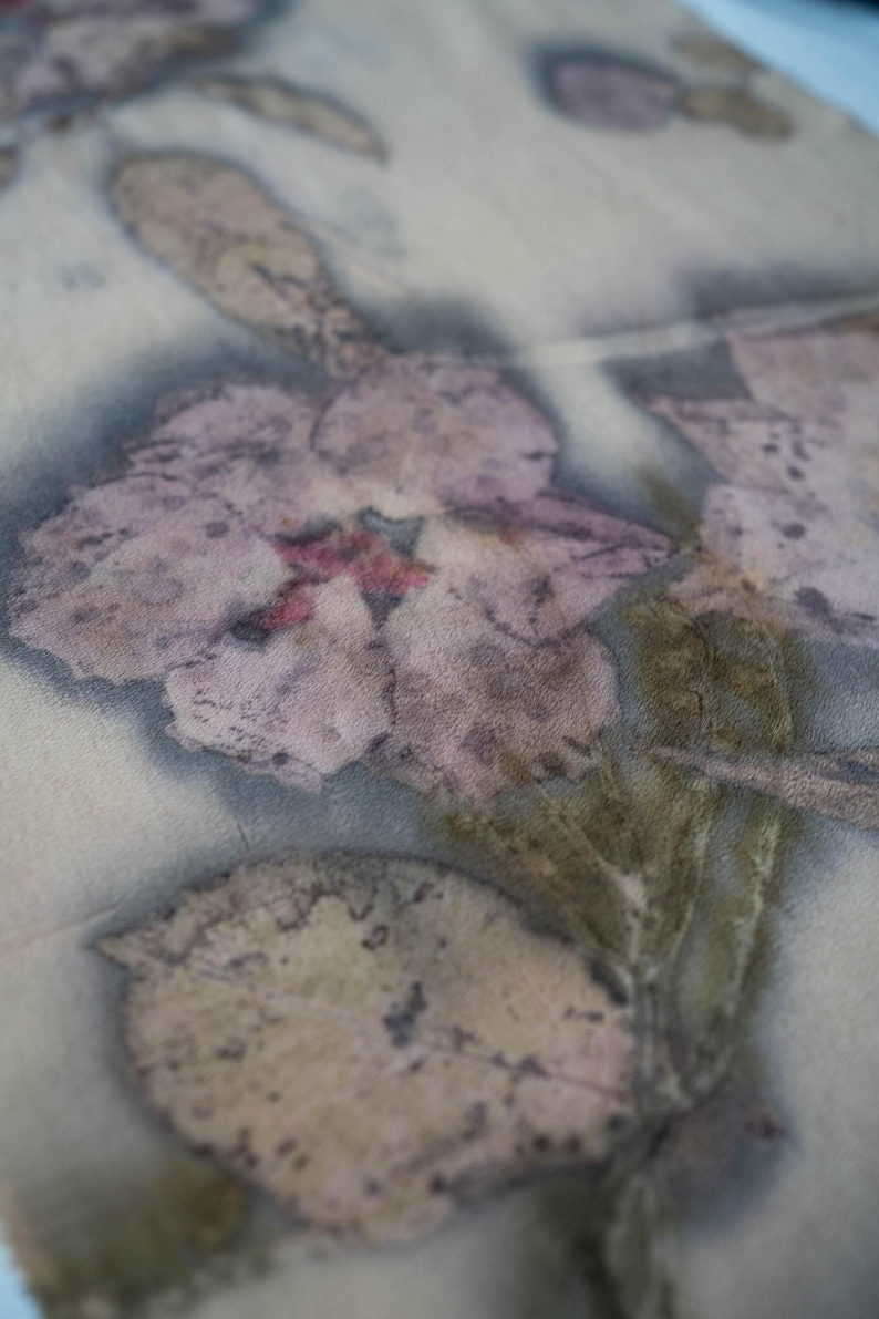May include: Close-up of a textile with a floral print. The design features a large, faded purple flower with a red center and several leaves in shades of brown and green. The fabric has a soft, muted color palette.