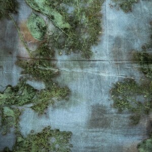 May include: A piece of fabric featuring a botanical print with green leaf and flower silhouettes against a muted blue and grey background. The textile displays organic, pressed plant patterns with visible veins and textures across the surface.