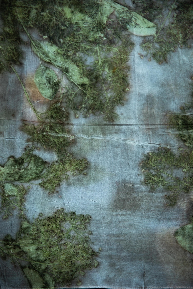 May include: A piece of fabric featuring a botanical print with green leaf and flower silhouettes against a muted blue and grey background. The textile displays organic, pressed plant patterns with visible veins and textures across the surface.