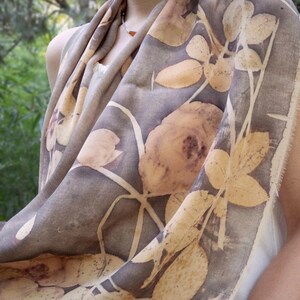 May include: A gray scarf with a botanical print featuring large, light brown rose and leaf designs. The scarf is draped, showcasing the detailed floral pattern and fringed edges. The fabric appears soft and lightweight.