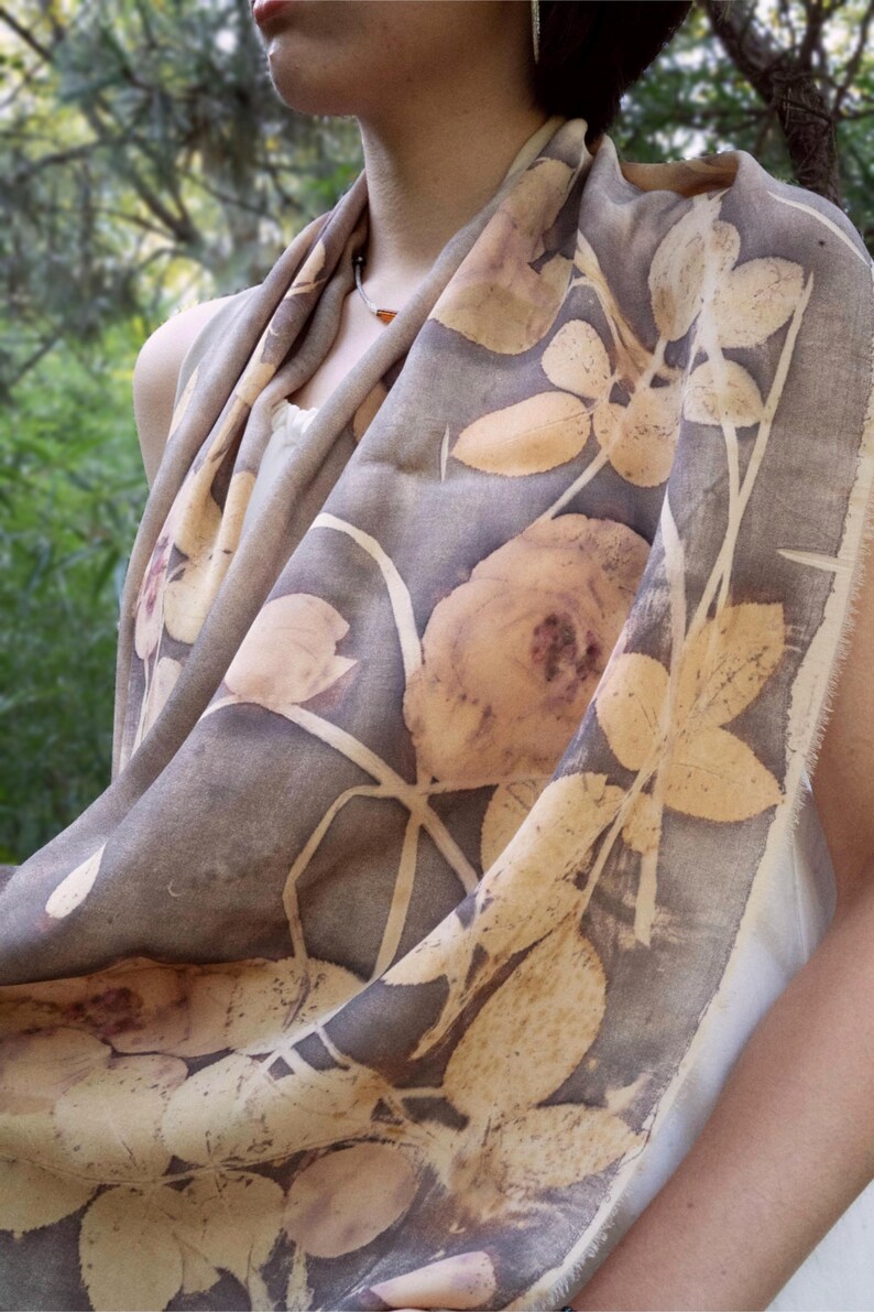 May include: A gray scarf with a botanical print featuring large, light brown rose and leaf designs. The scarf is draped, showcasing the detailed floral pattern and fringed edges. The fabric appears soft and lightweight.