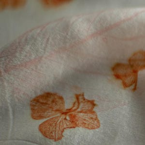 May include: A white fabric featuring a repeating botanical print of orange flower blossoms and faint leaf veins. The material is draped to create soft folds, highlighting the texture of the textile and the detailed orange floral patterns.