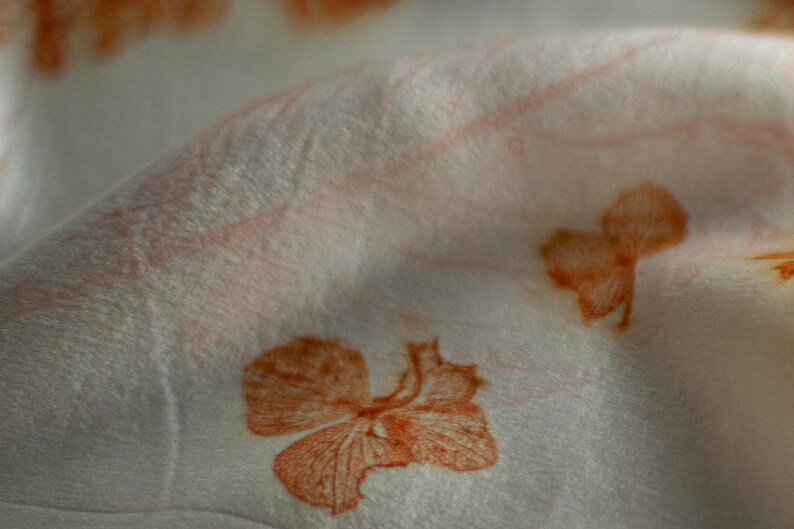 May include: A white fabric featuring a repeating botanical print of orange flower blossoms and faint leaf veins. The material is draped to create soft folds, highlighting the texture of the textile and the detailed orange floral patterns.