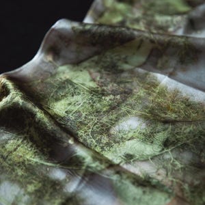 May include: A lightweight fabric scarf featuring a botanical print with intricate green leaf and fern patterns. The material has a soft, draped texture with visible organic veining and earthy tones layered over a pale, neutral base color.