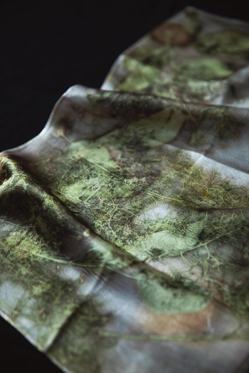 May include: A lightweight fabric scarf featuring a botanical print with intricate green leaf and fern patterns. The material has a soft, draped texture with visible organic veining and earthy tones layered over a pale, neutral base color.