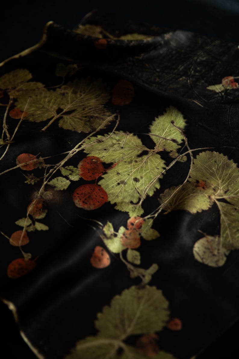 May include: A black fabric featuring a botanical print of green serrated leaves and small, round, reddish-brown berries. The design displays a natural, organic arrangement of foliage and fruit scattered across the dark, smooth surface of the textile.