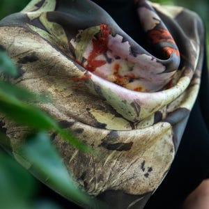 May include: A square scarf featuring a botanical print with earthy tones of brown, beige, and muted green, accented by vibrant orange and deep red leaf patterns. The fabric displays intricate, detailed textures of foliage and organic plant imprints.